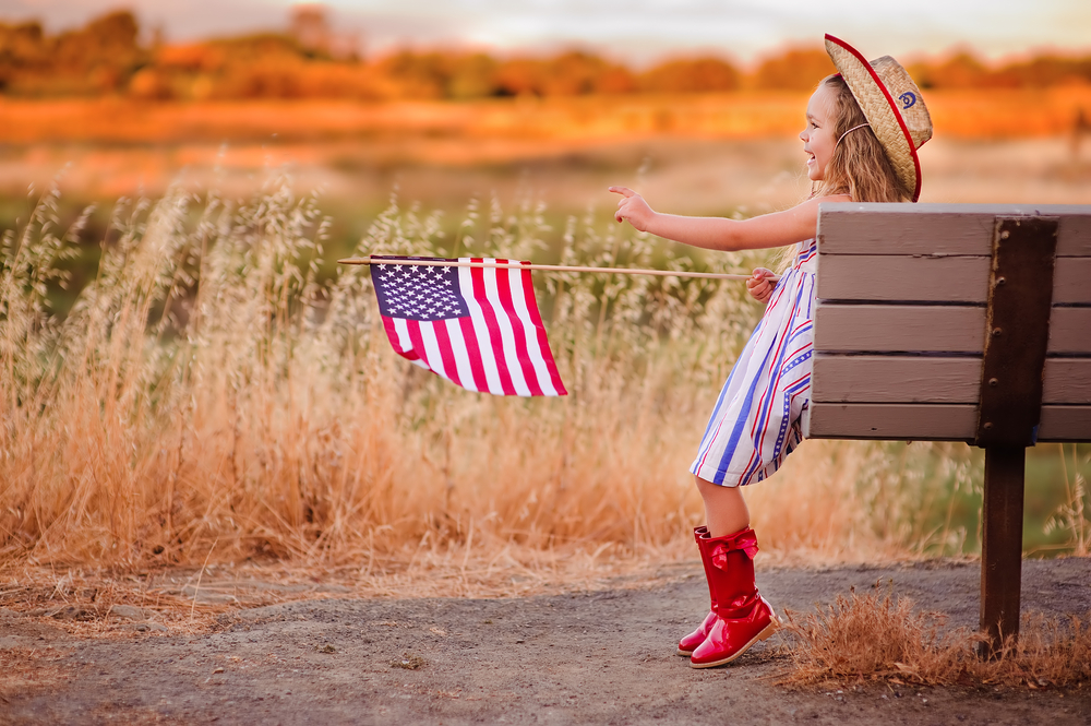 Young girl sitting on a bench in red boots and holding an American Flag