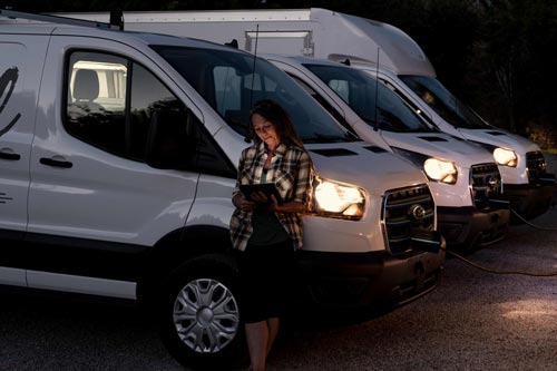 A woman getting work done while a commercial fleet of 2025 Ford E-Transit Vans charge