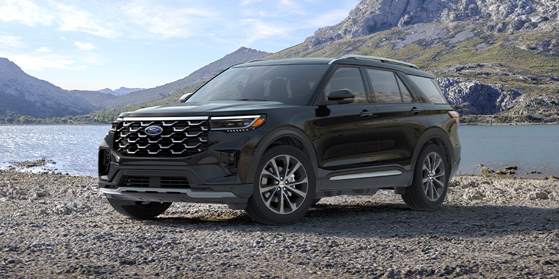 Front driver's side view of a black 2025 Ford Explorer parked next to a lake with hills behind and a slightly cloudy blue sky overhead